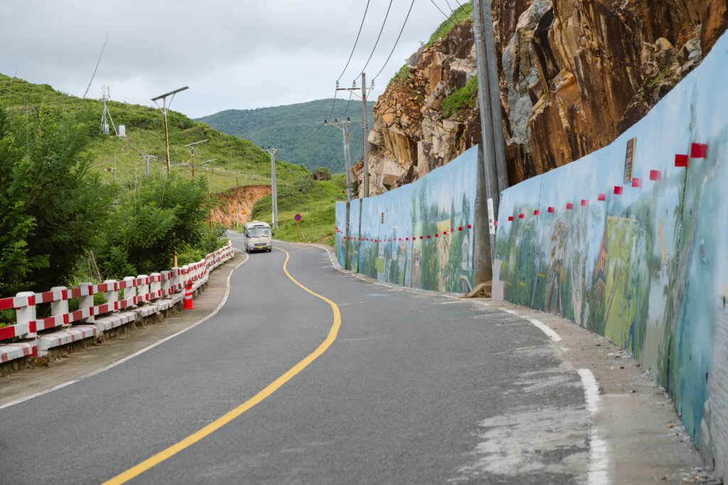 A scenic coastal road winding past a newly painted, extensive concrete wall mural.