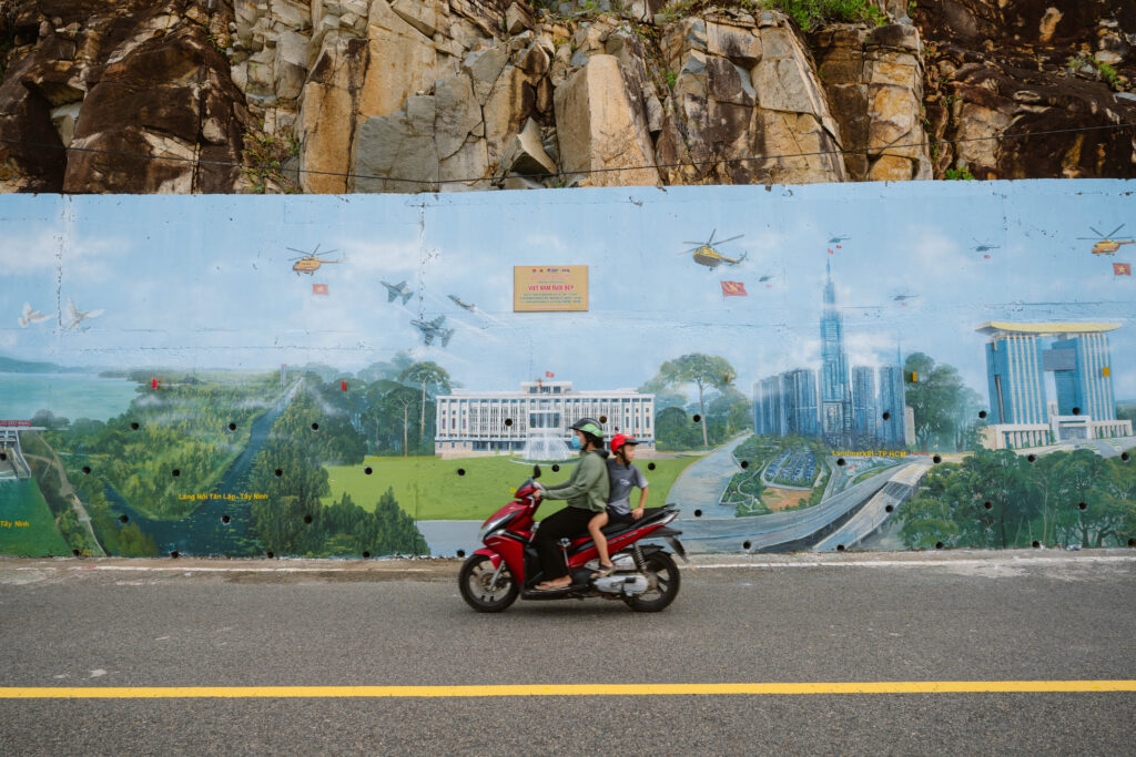 Two people riding a red scooter past a vibrant mural depicting landmark Vietnamese architecture.