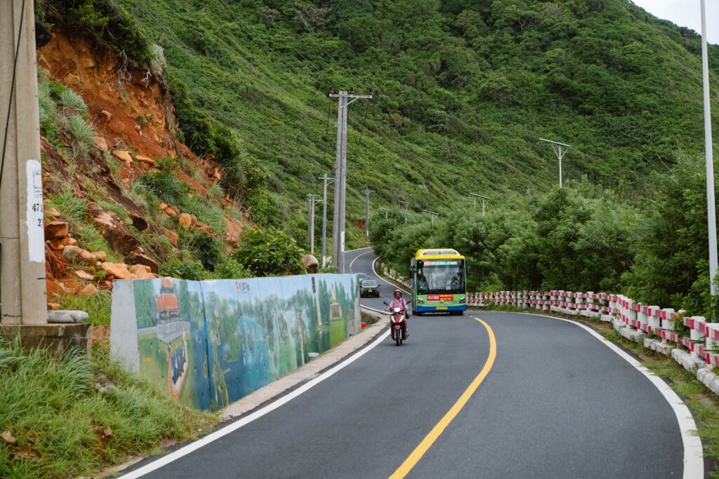 A bus and scooter navigating a road bordered by a colourful community mural against a hillside.