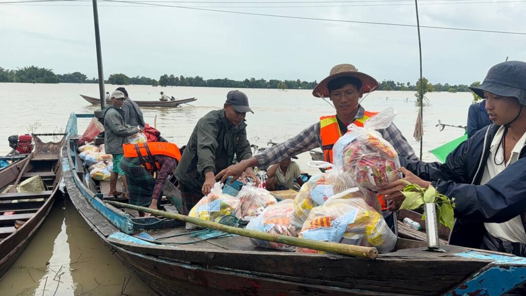 Volunteers in life jackets loading bags of food rations onto a wooden boat in floodwaters.