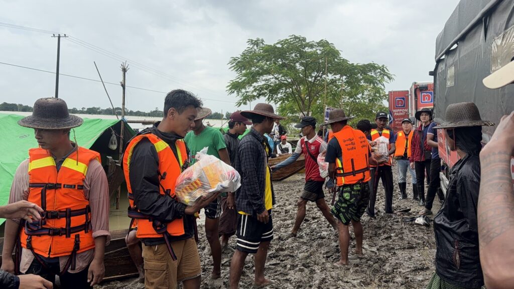 A line of volunteers passing bags of essential supplies across muddy ground towards a boat.